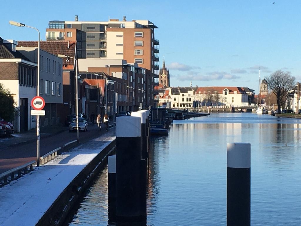 Foto van de oever vab de Schie met links bebouwing, rechts het water en op de achtergrond de Delftse binnenstad.